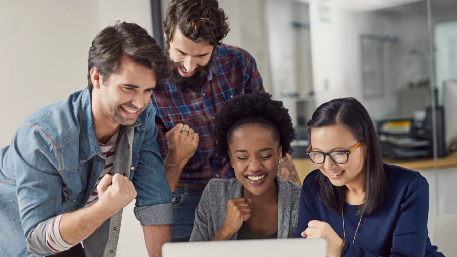 Foto de quatro colegas reunidos em volta de um notebook, sorrindo e comemorando com os punhos cerrados