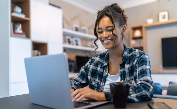 Foto de uma mulher de camisa xadrez sorrindo enquanto digita no notebook em casa, com uma caneca ao lado