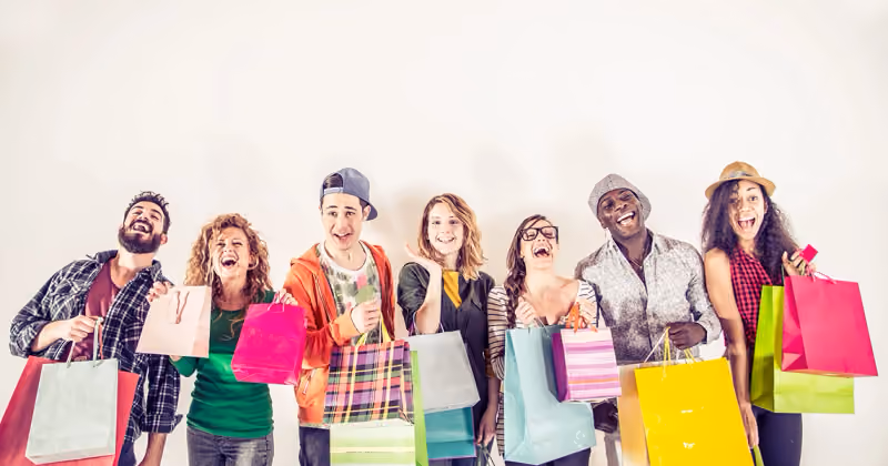 Foto de sete jovens sorrindo lado a lado contra parede branca, segurando várias sacolas de compras coloridas e um cartão rosa